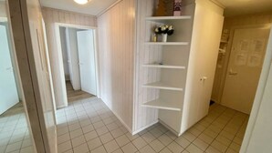 En gang med hvide flisegulve, en træskab med hylder og en dør med et skilt.
