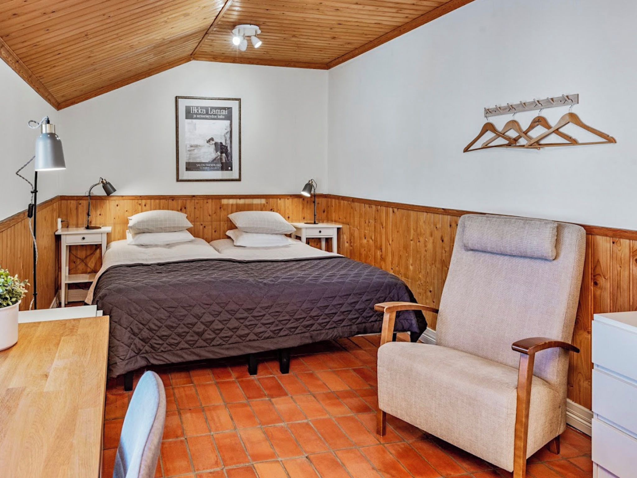 Une chambre avec un plafond en bois, un lit recouvert d’une courtepointe, une chaise, un bureau et un porte-manteau fixé au mur.