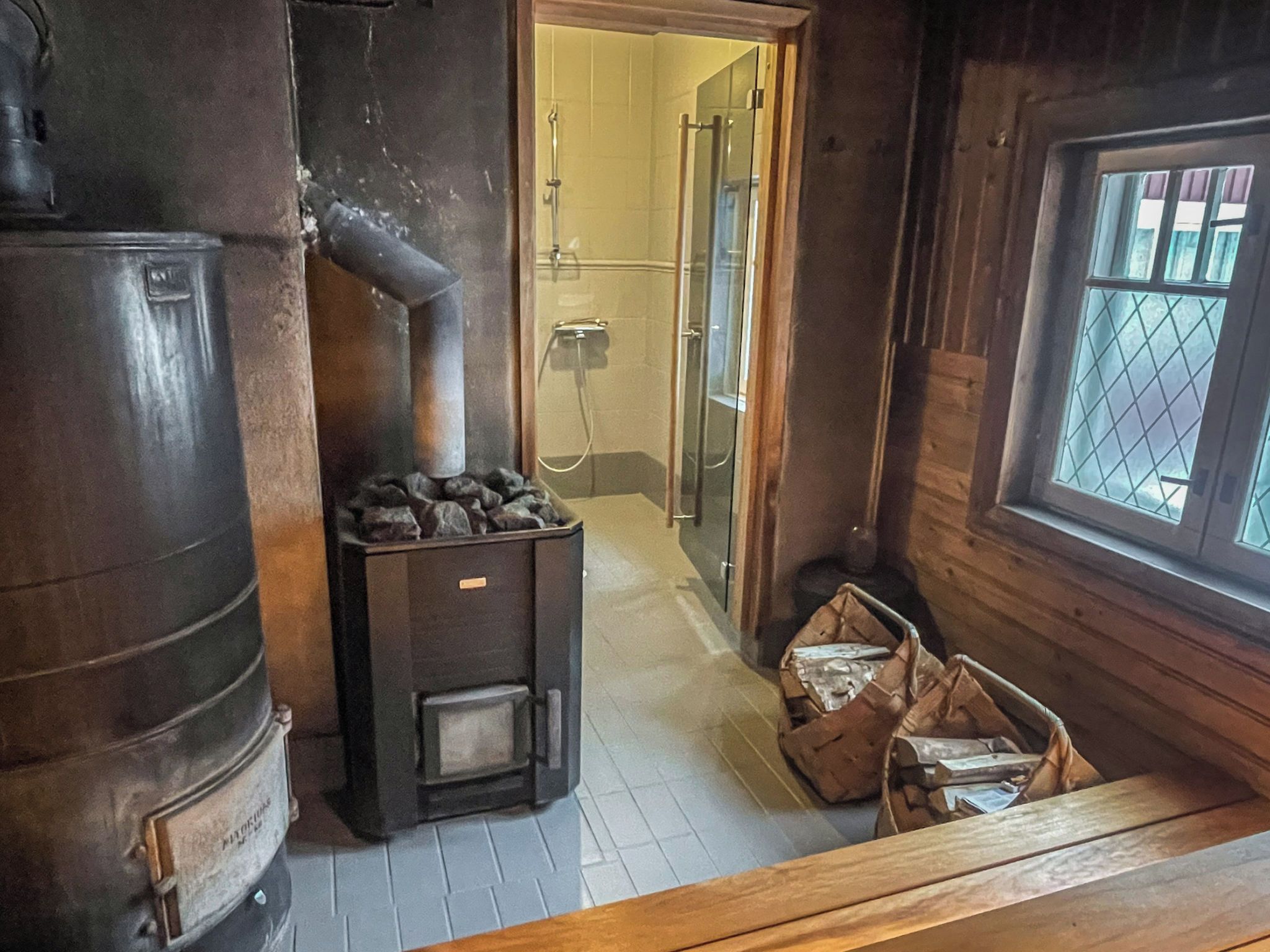 A rustic cabin interior with a wood-burning stove, a shower area, and a window with a view.