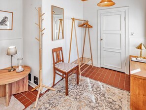 Una habitación con una silla de madera, un perchero, un abrigo colgado, una puerta, una lámpara y un cuadro en la pared.
