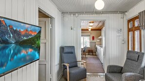 A cozy interior with a large TV displaying a scenic mountain lake, two grey armchairs, a wooden table, and a kitchen area in the background. - Punatulkku by Interhome (Somero)