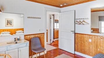 A room with a wooden ceiling, a white door, a mirror, a bed, a chair, and a bathroom with a sink and mirror.