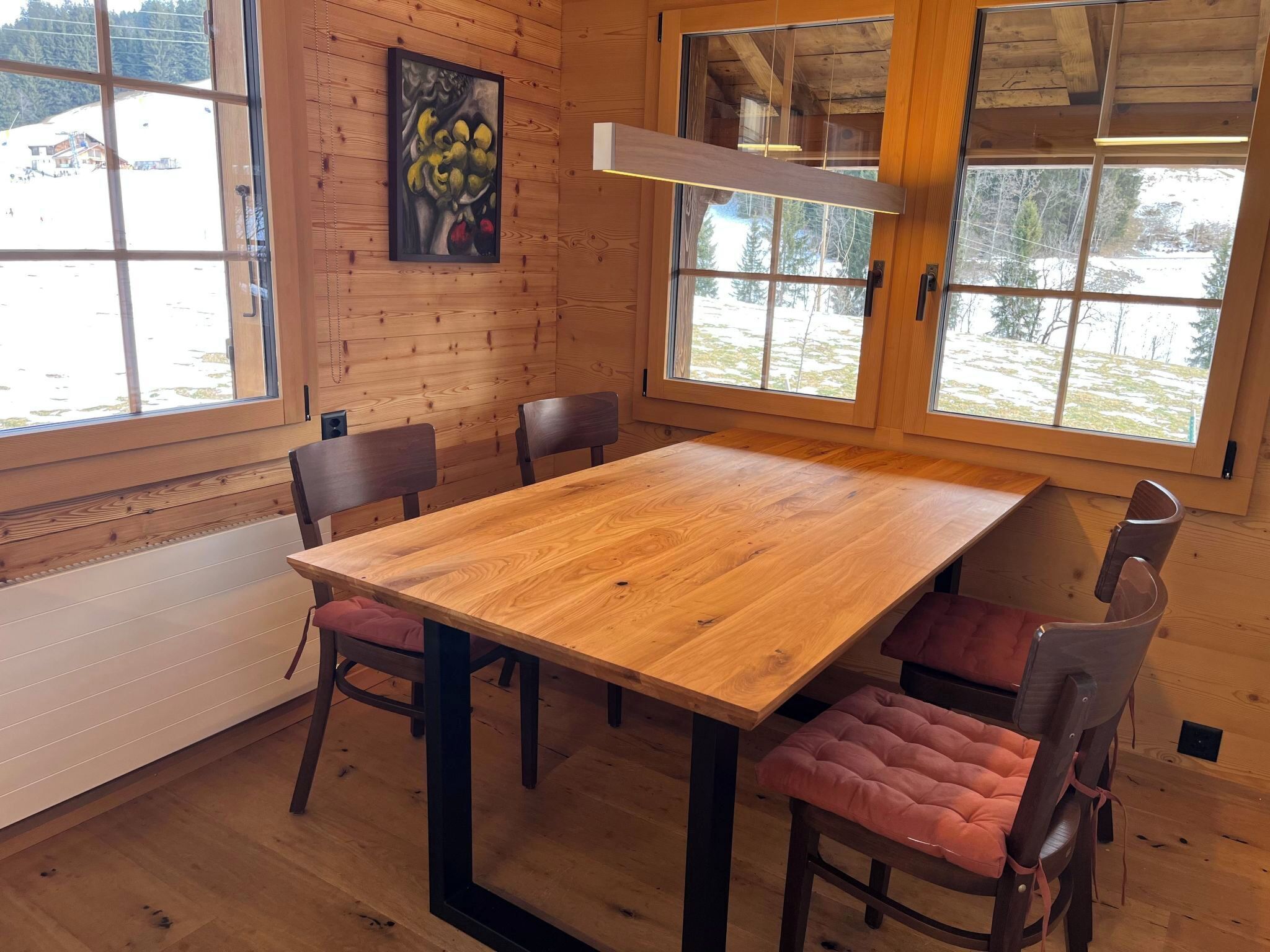 Een houten interieur met een grote tafel en stoelen, grote ramen en een schilderij aan de muur.