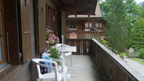 Un balcón de madera con sillas de plástico blanco y una pequeña mesa, con vistas a un jardín con árboles y un edificio al fondo.