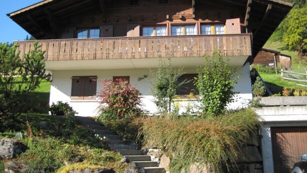 Un chalet de dos pisos con un balcón de madera, escalones de piedra que conducen a la entrada y un jardín con arbustos y una planta en flor.