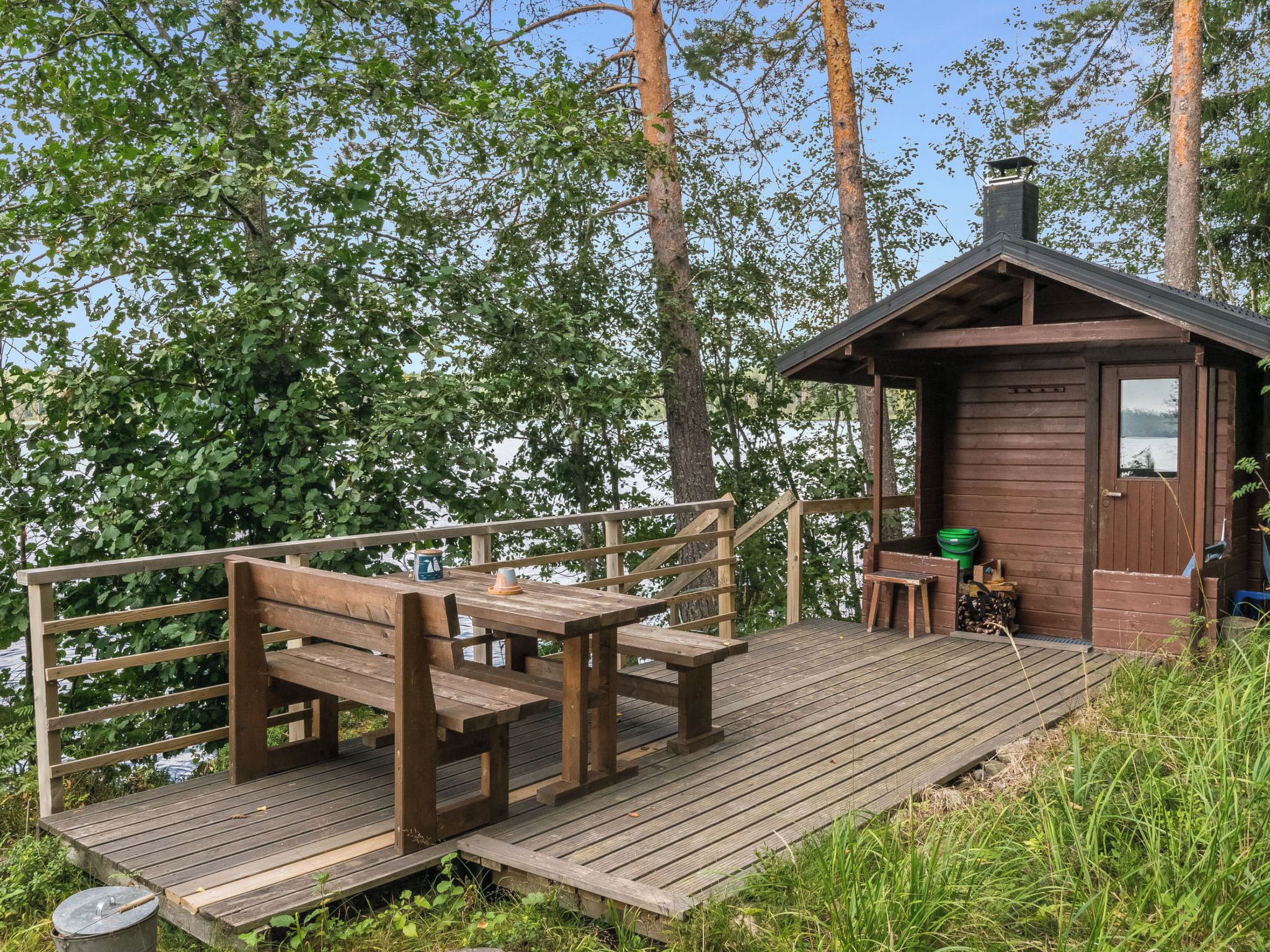 Een houten terras met banken en een tafel, een klein huisje met schoorsteen, en uitzicht op een meer omringd door bomen.