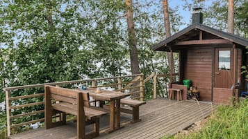 Een houten terras met banken en een tafel, een klein huisje met schoorsteen, en uitzicht op een meer omringd door bomen.