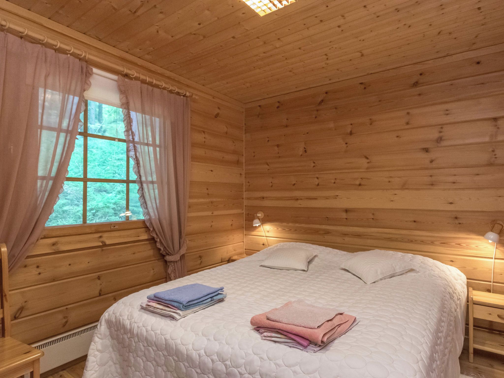 A wooden cabin room with a bed, two bedside lamps, a window with curtains, and a radiator.