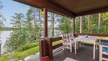 A covered outdoor deck with a table and chairs, overlooking a body of water and surrounded by trees.