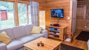 A cozy cabin interior with a sofa, a wooden table, a TV mounted on a wall, and a window with curtains.