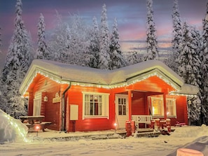 Ein mit Schnee bedecktes, rotes Blockhaus mit einer Veranda, Bänken und einem kleinen Tisch.