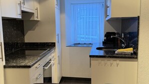A compact kitchen with white cabinets, a black countertop, and a window with sheer curtains. There is a small island with a sink and a stove. - Mier Gfallt's by Interhome (Saas-Fee)