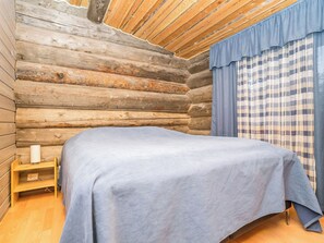 Un dormitorio de cabaña de madera con cama, una mesita de noche pequeña de madera y una ventana con cortinas a cuadros.