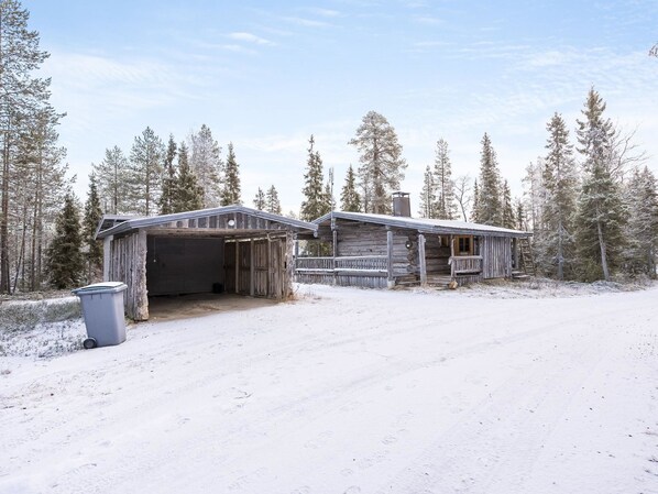 A rustic cabin with a garage and a porch, surrounded by snow-covered trees.
