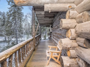 Una terraza de madera que lleva a una cabaña de troncos con barandilla metálica, rodeada de árboles cubiertos de nieve.