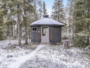 Una cabaña pequeña de madera con puerta y ventanas, rodeada de suelo y árboles cubiertos de nieve.
