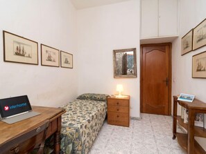 Un dormitorio con cama, un escritorio de madera con una computadora portátil, una mesita de noche con lámpara y una puerta de madera.
