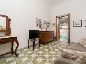 Une pièce avec un téléviseur posé sur un meuble, une commode en bois, un miroir et un canapé avec des coussins. Des tableaux encadrés ornent le mur et, à travers une porte vitrée, on aperçoit une piscine.