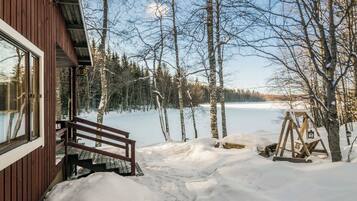 Una cabaña de madera con un banco en el exterior, rodeada de árboles cubiertos de nieve y un lago congelado.