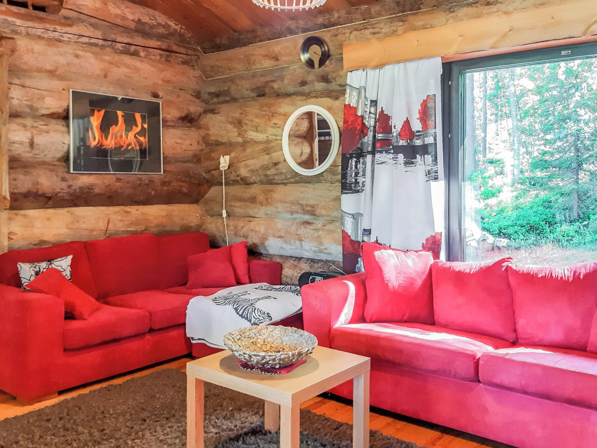 Interior de una cabaña de troncos con dos sofás rojos, una mesita con un cuenco, un reloj de pared y un ventanal con vistas a follaje.