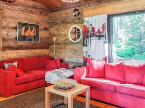 A log cabin interior with two red sofas, a small table with a bowl, a wall clock, and a window with a view of greenery.