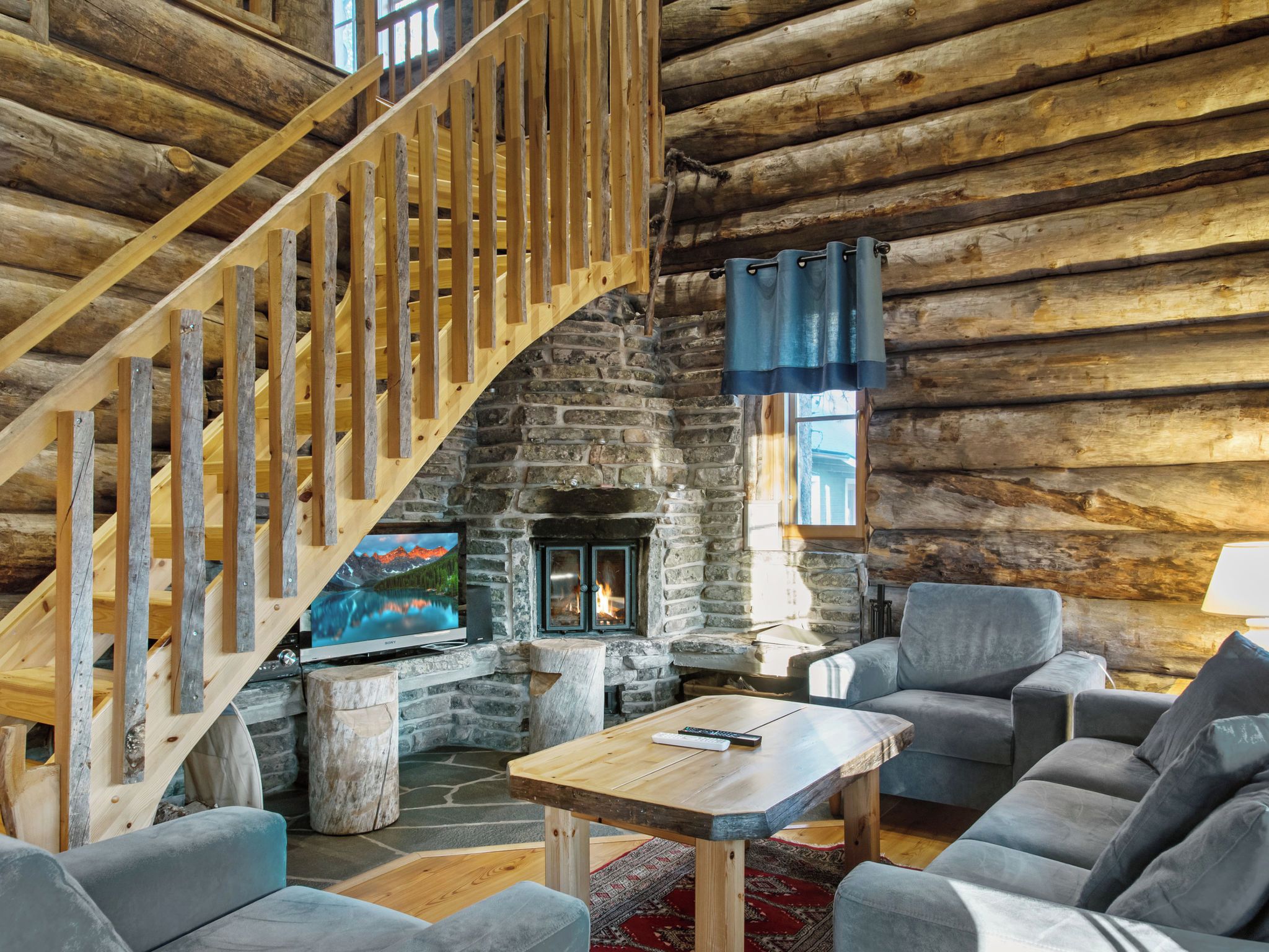 A log cabin interior with a stone fireplace, a wooden staircase, and a television.