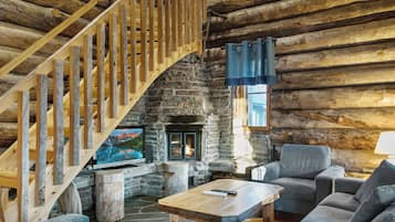 A log cabin interior with a stone fireplace, a wooden staircase, and a television.