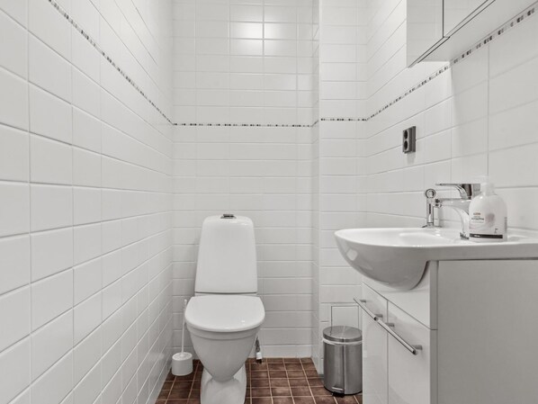 A small, modern bathroom with white tiles, a white toilet, a sink, and a trash can.