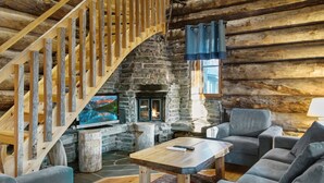 Interior de una cabaña de troncos con chimenea de piedra, una escalera de madera y una zona de estar acogedora con televisor de pantalla plana.
