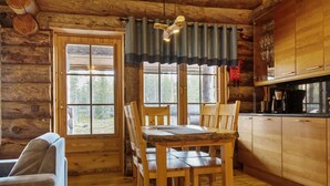 Interior rústico de una cabaña con una mesa y sillas de madera, una zona de cocina con armarios de madera, y ventanas amplias que dan a un bosque circundante.