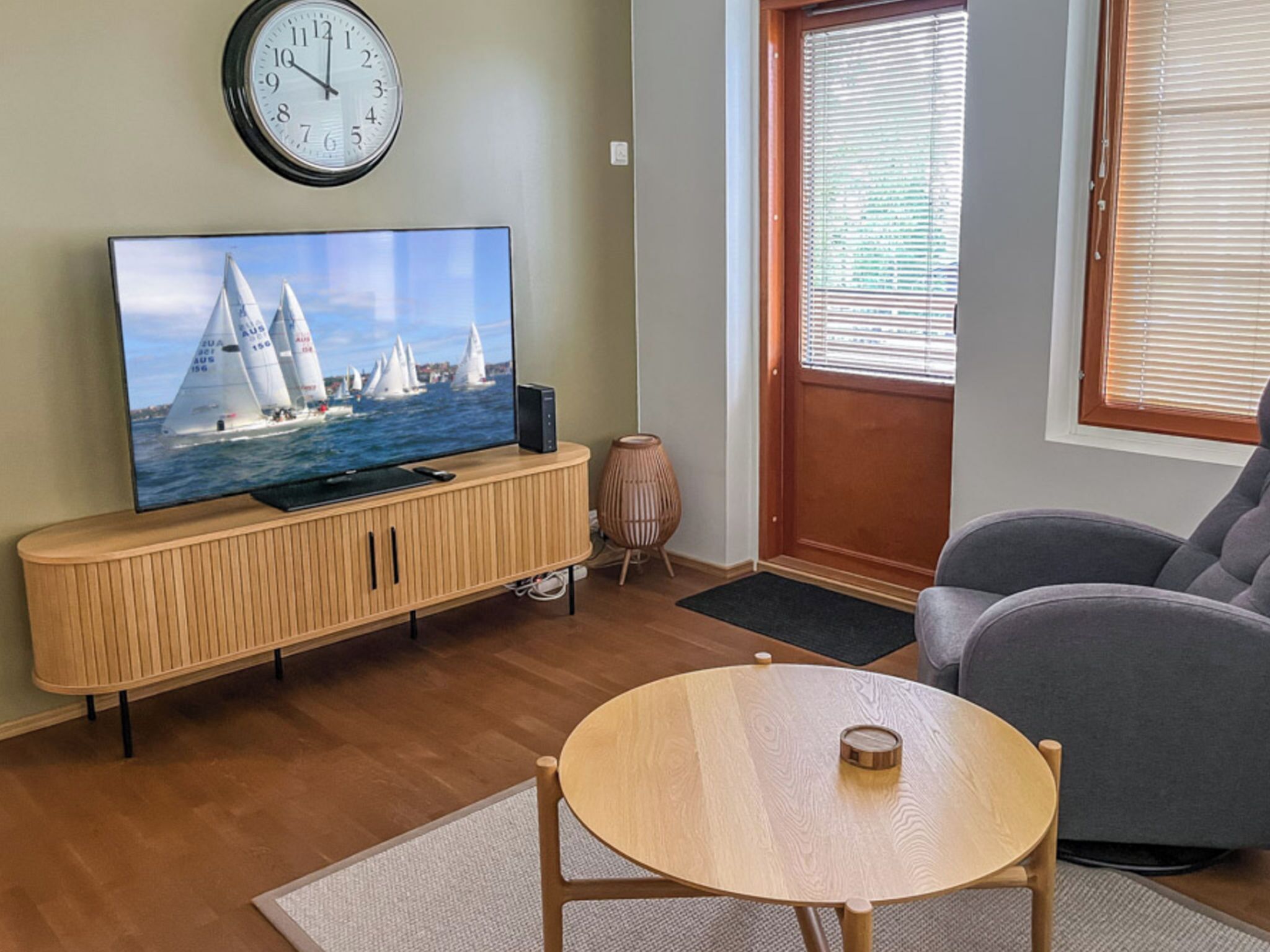 Ein Wohnzimmer mit Fernseher auf einer Holzkommode, einem runden Tisch, einem grauen Sofa, einer Wanduhr und einer Tür mit Jalousien.