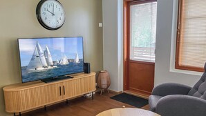Uma sala de estar com televisão sobre um móvel de madeira, uma mesa redonda, um sofá cinzento, um relógio na parede e uma porta com persianas.