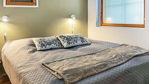 Uma cama bem arrumada com colcha acolchoada e duas almofadas decorativas, um abajur e uma janela com persianas.