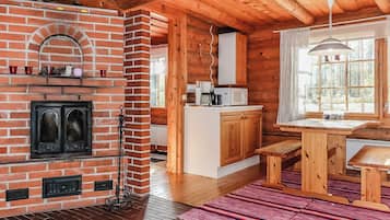 A cozy cabin interior with a brick fireplace, wooden beams, a kitchen area, and a large window.