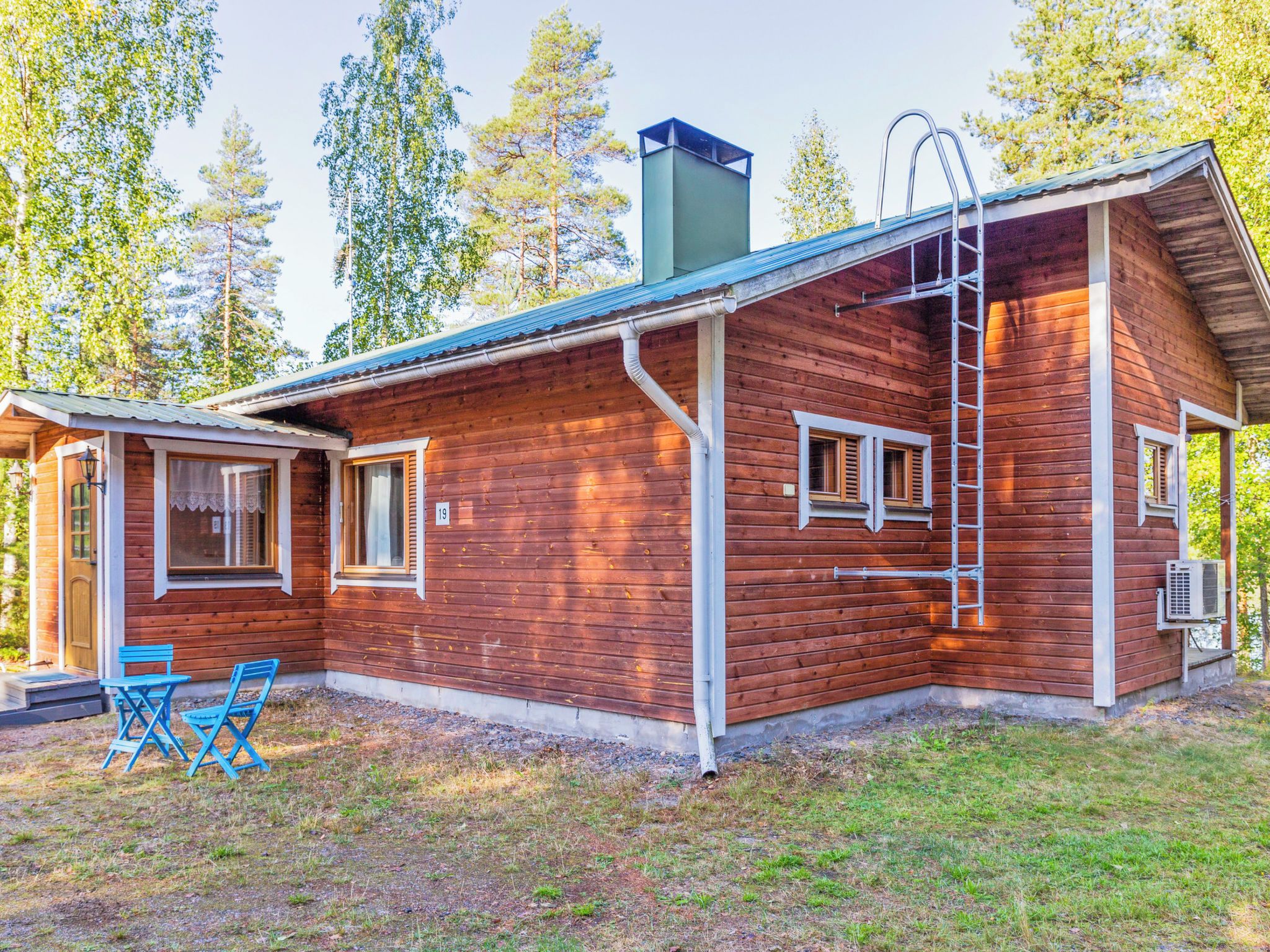 Puinen mökki, jossa on savupiippu, sinisillä tuoleilla varustettu kuisti ja mökin sivussa oleva tikapuu.