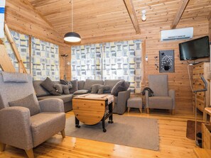 Ein gemütliches Cabin-Interieur mit einer Couch, Sesseln, einem Holztisch, einem Fernseher und einer gemusterten Fensterdekoration.