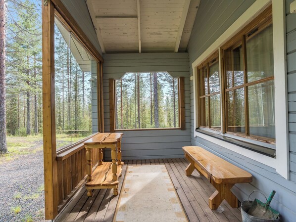 Eine überdachte Veranda mit Holzbänken und -tischen, großen Fenstern und Blick auf einen Wald.