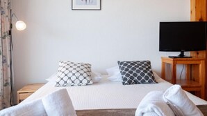 Ein Schlafzimmer mit einem Bett, zwei Kissen mit Muster, einem gerahmten abstrakten Kunstwerk, einem Fernseher auf einem Holzständer und einer Stehlampe.