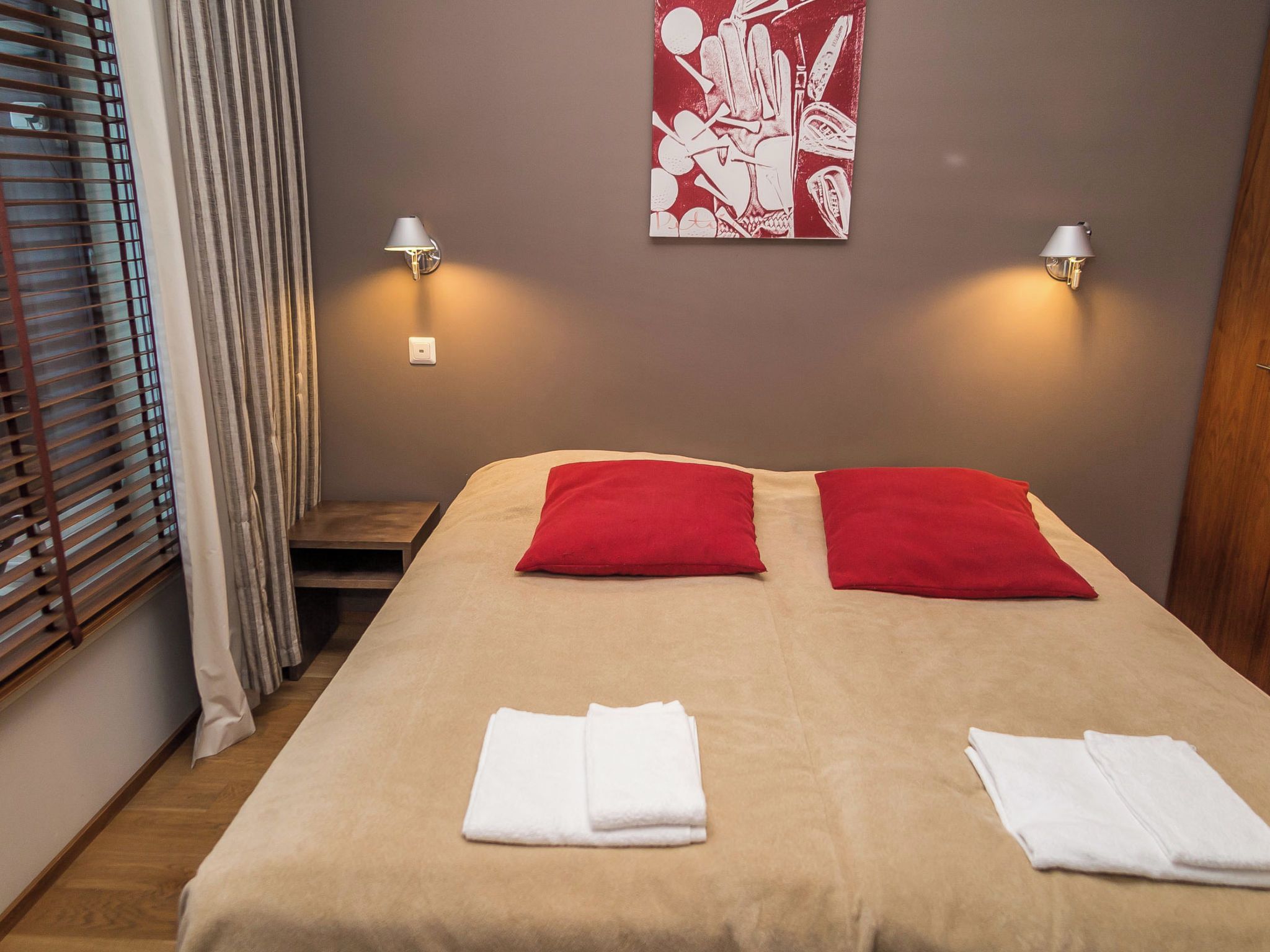 Une chambre d’hôtel avec un lit, deux oreillers, deux lampes fixées au mur, une fenêtre avec des stores et un tableau accroché au mur.