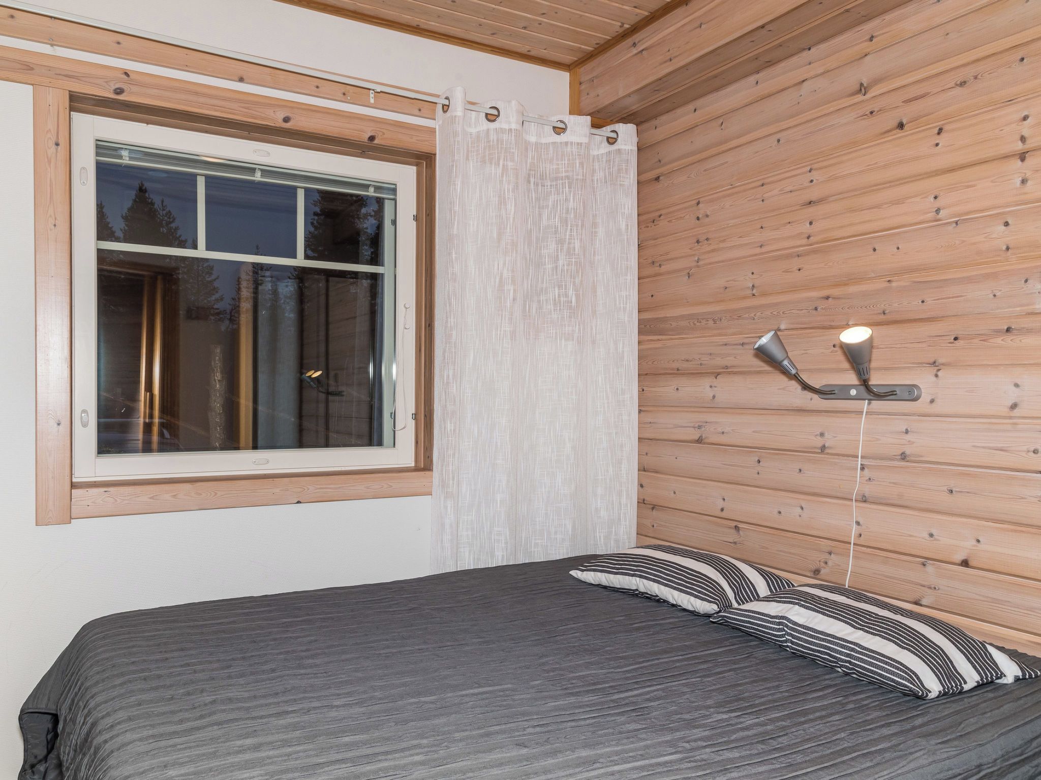 A room with a bed, a window with curtains, and a wall-mounted lamp.