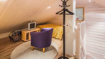 Um quarto de sótão pequeno e acolhedor, com teto inclinado, uma cama com manta amarela, uma cadeira roxa, uma pequena mesa de madeira com uma televisão e um abajur.