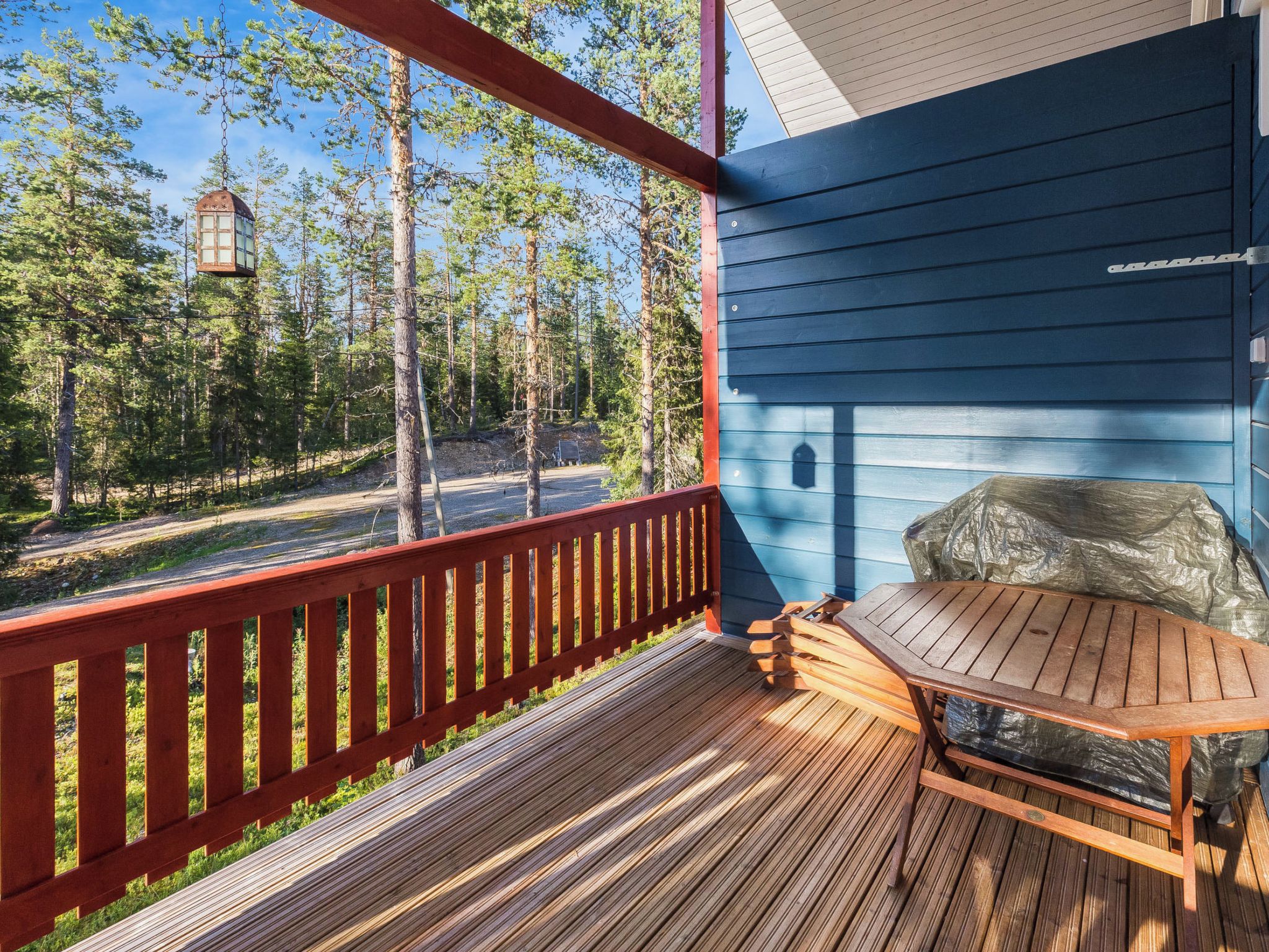 Um deck de madeira com guarda-corpo vermelho, uma mesa e um edifício azul com detalhe em pedra.