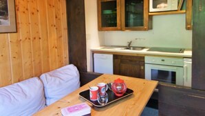 A cozy room with a wooden ceiling, a sofa, a dining table with cups and a tray, and a kitchen area with a microwave and sink. - Grand Roc by Interhome (Argentière)