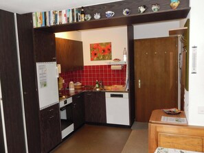 A compact kitchen with a refrigerator, oven, and dishwasher, a red tiled backsplash, and a small shelf with decorative items. - Sörenweg 4 Appartement/Fewo 2-Bettwohnung by Interhome (Engelberg)