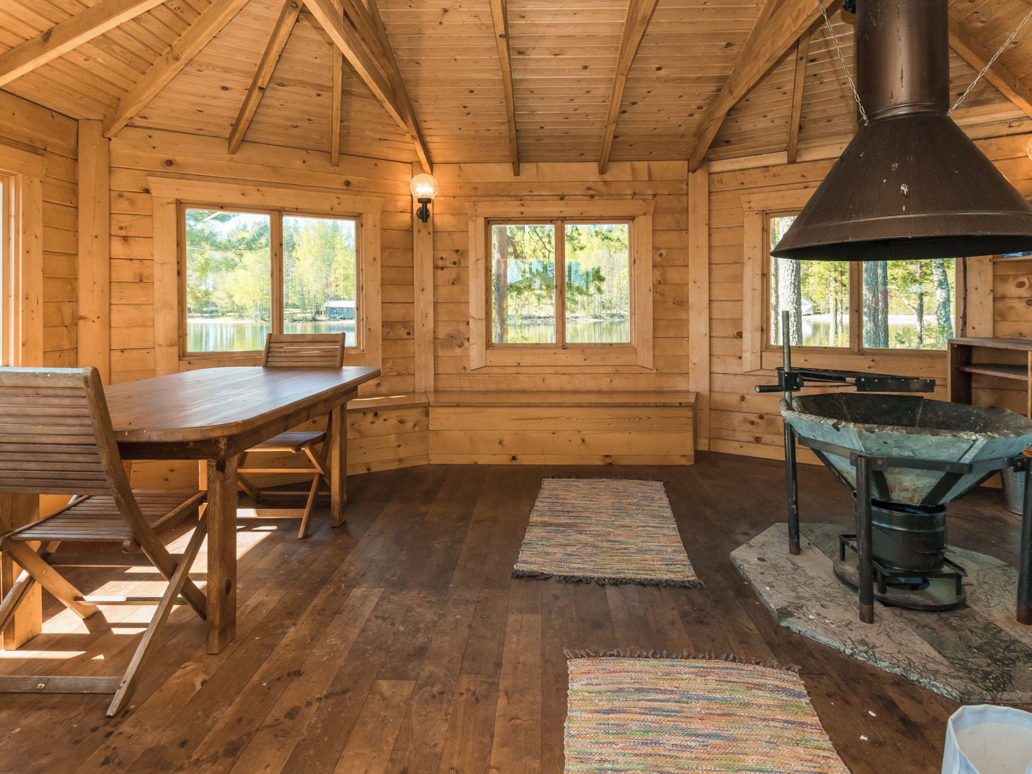 A wooden cabin interior with a dining table, benches, a large chimney, and windows overlooking a natural landscape.