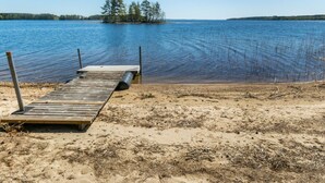 A wooden dock extending into a calm lake with a sandy shore and a small island with trees in the distance. - Villa pyöriäinen by Interhome (Kerimäki)