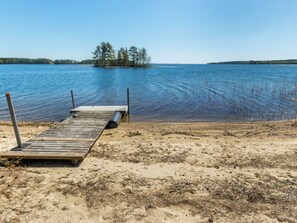 A wooden dock extending into a calm lake with a sandy shore and a small island with trees in the distance. - Villa pyöriäinen by Interhome (Kerimäki)
