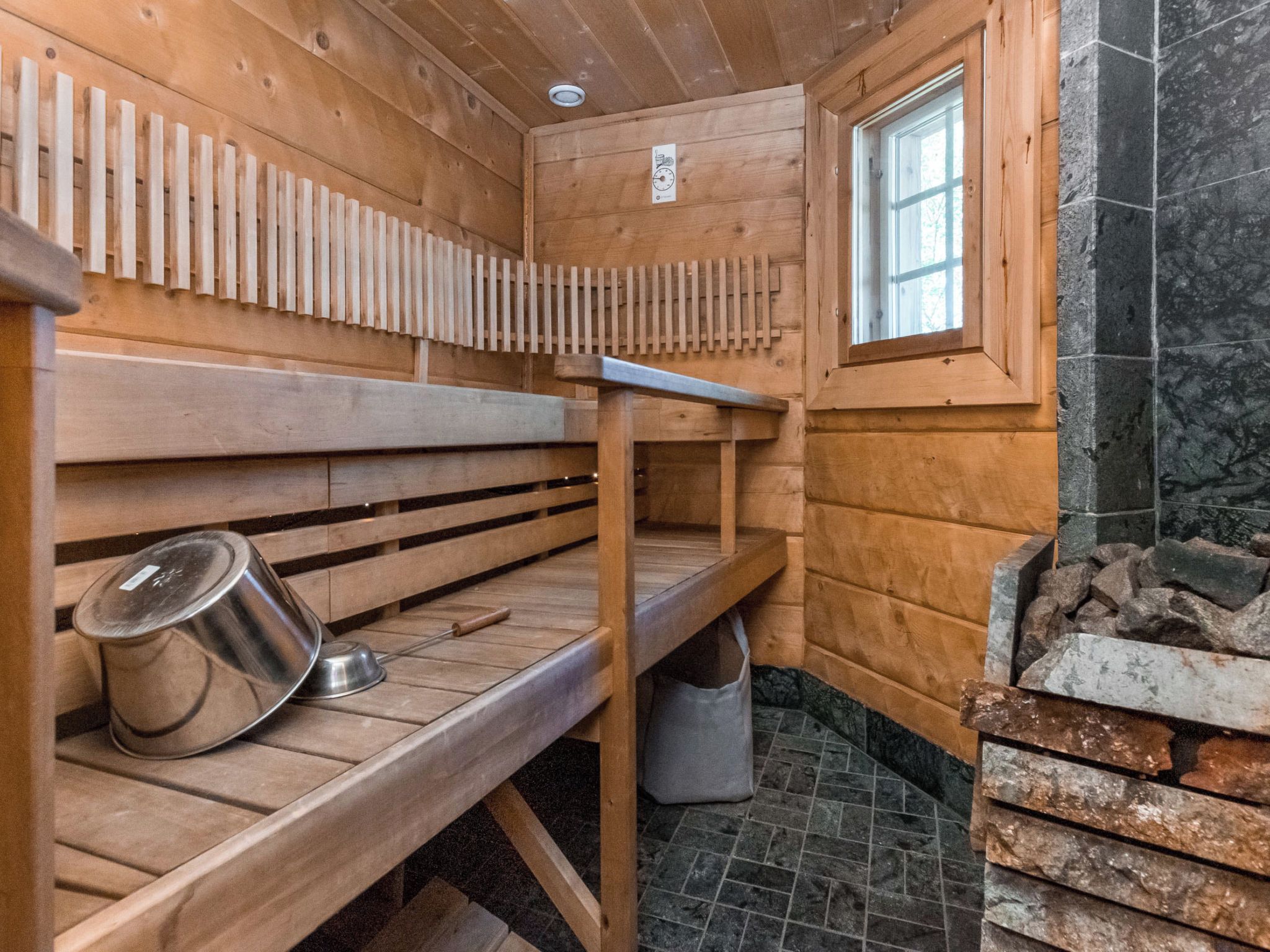 A sauna room with wooden benches, a metal pot, a window, and a stone wall.