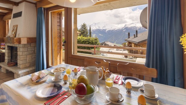 A dining table set with breakfast items, a fireplace, and a view of mountains through a window. - Mogador 13 by Interhome (Verbier)