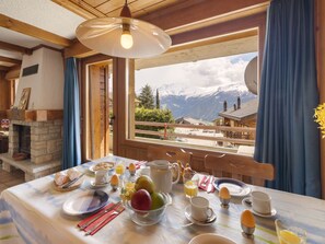 Une table à manger dressée avec des aliments pour le petit-déjeuner, une cheminée et une vue sur les montagnes par la fenêtre.